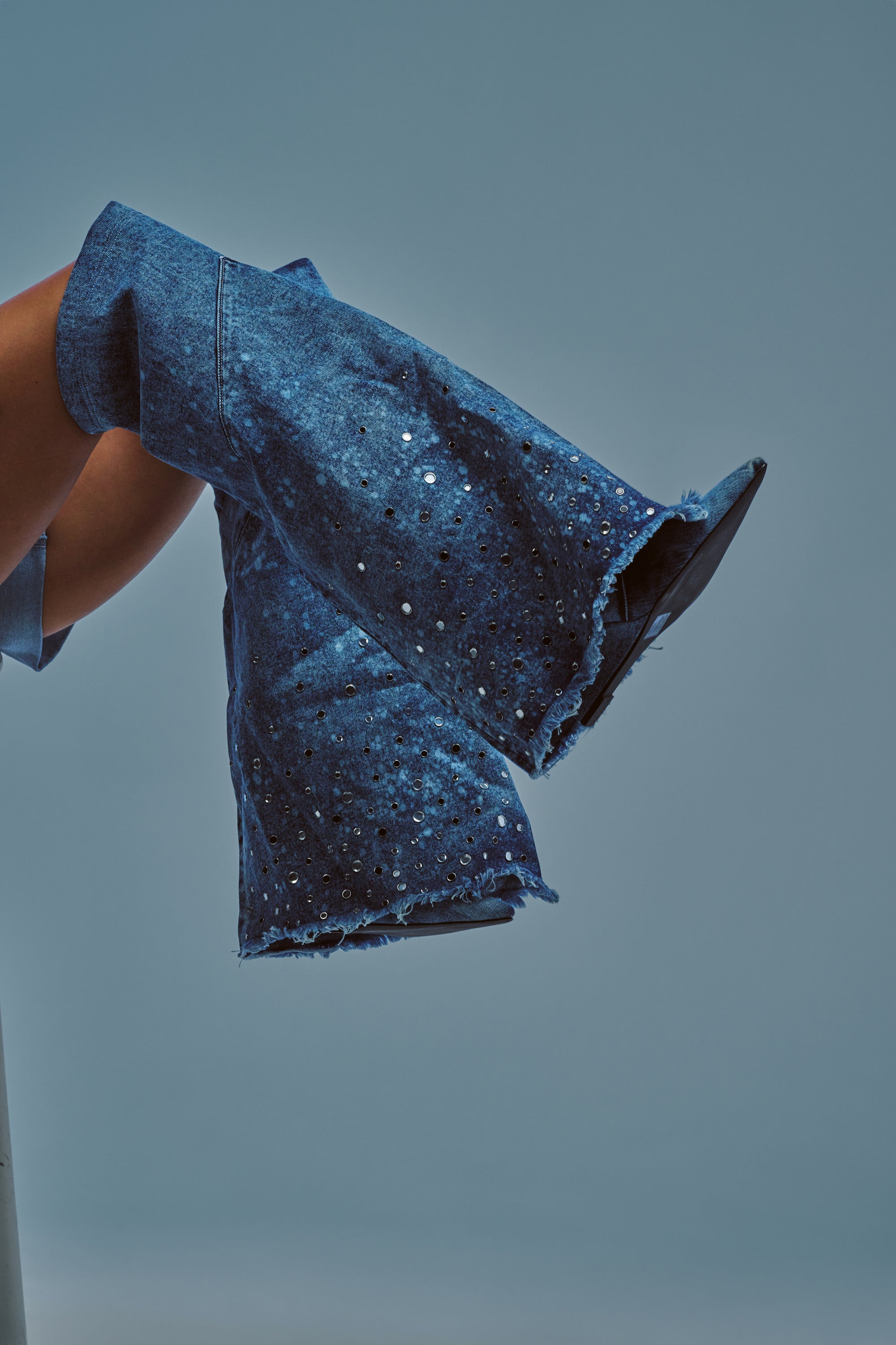 A person’s legs are crossed, clad in flared blue denim pants with frayed hems and sparkling rhinestone details, effortlessly styled with the AKIRA WHYNOT DENIM RHINESTONE EMBELLISHED WEDGE BOOT, all set against a plain blue-gray background.