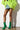 A person wearing a bright green fluffy coat, a denim miniskirt, and AZALEA WANG WILBER BOOTIE WITH STRETCH NEOPRENE IN GREEN featuring metallic stiletto heels stands against a wall. The natural light casts a shadow on the white background.