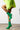 A person wearing a vivid green fuzzy sweater and the AZALEA WANG Wilber Bootie with stretch neoprene, featuring metallic stiletto heels, is captured mid-step while lifting one leg. The plain white wall in the background and the neutral-toned flooring complement the scene.