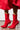 A person is wearing the AZALEA WANG WILBER Bootie with Stretch Neoprene in Red, featuring a bright red design with pointy toes and stiletto heels. These unique booties are perfectly paired with a red dress in a simple indoor setting, complemented by a concrete floor.