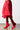 A person is wearing a voluminous red skirt paired with the eye-catching AZALEA WANG Wilber booties, which feature pointy toes, stiletto heels, and red stretch neoprene. The scene captures the lower half of the body against a plain light background.