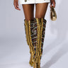 Dressed in a white dress adorned with detailed floral embroidery, the individual complements their look with the AZALEA WANG WILDA MULTI STILETTO BOOT. These tall, decorative boots boast vibrant patterns and gold accents, enhanced by a faux suede finish for an elegant touch, accompanied by a small matching gold accessory.