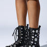 A person wears knee-length denim shorts and the AZALEA WANG Wilhelmina black embellished booties, which are adorned with large, shiny rhinestones. These lace-up booties feature a chunky heel and a buckle strap, set against a plain white background.