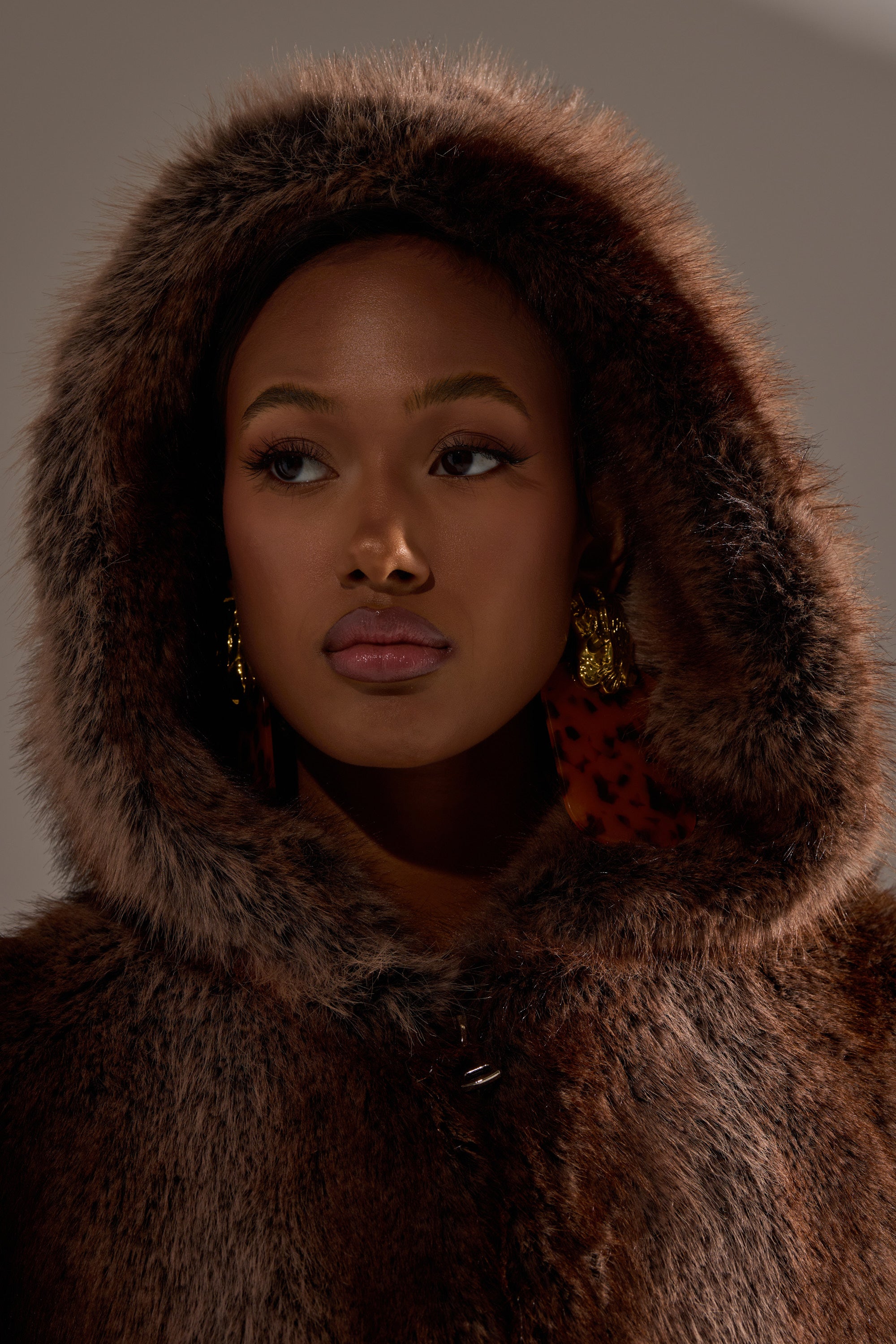 A woman with gold earrings and natural makeup gazes sideways in a neutral, softly lit setting, wearing the NATURAL INSTINCTS HOODED FUR BOMBER—an oversized winter coat with a large hood that frames her face.