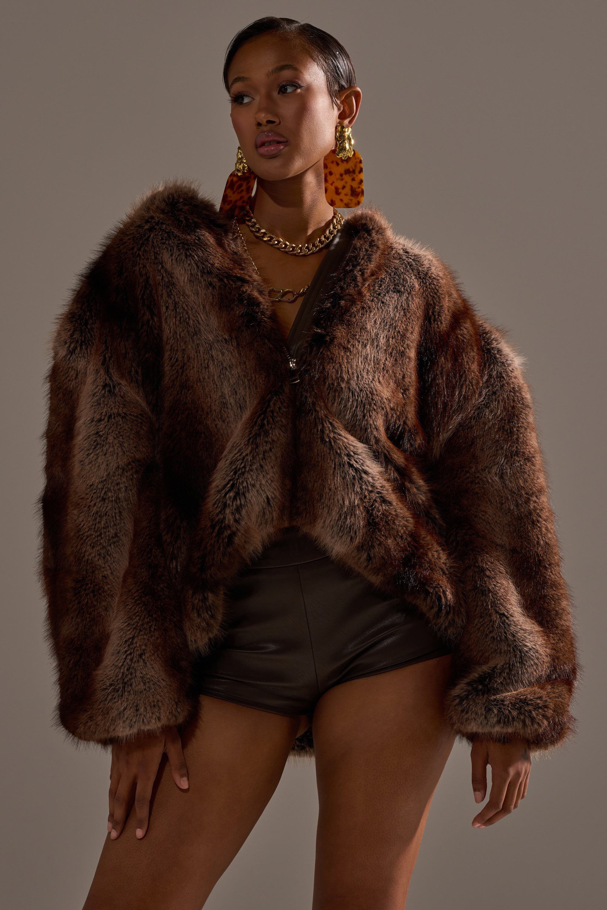 A woman models the NATURAL INSTINCTS HOODED FUR BOMBER with a gold chain necklace, bold tortoiseshell earrings, and dark high-waisted shorts against a plain gray background.