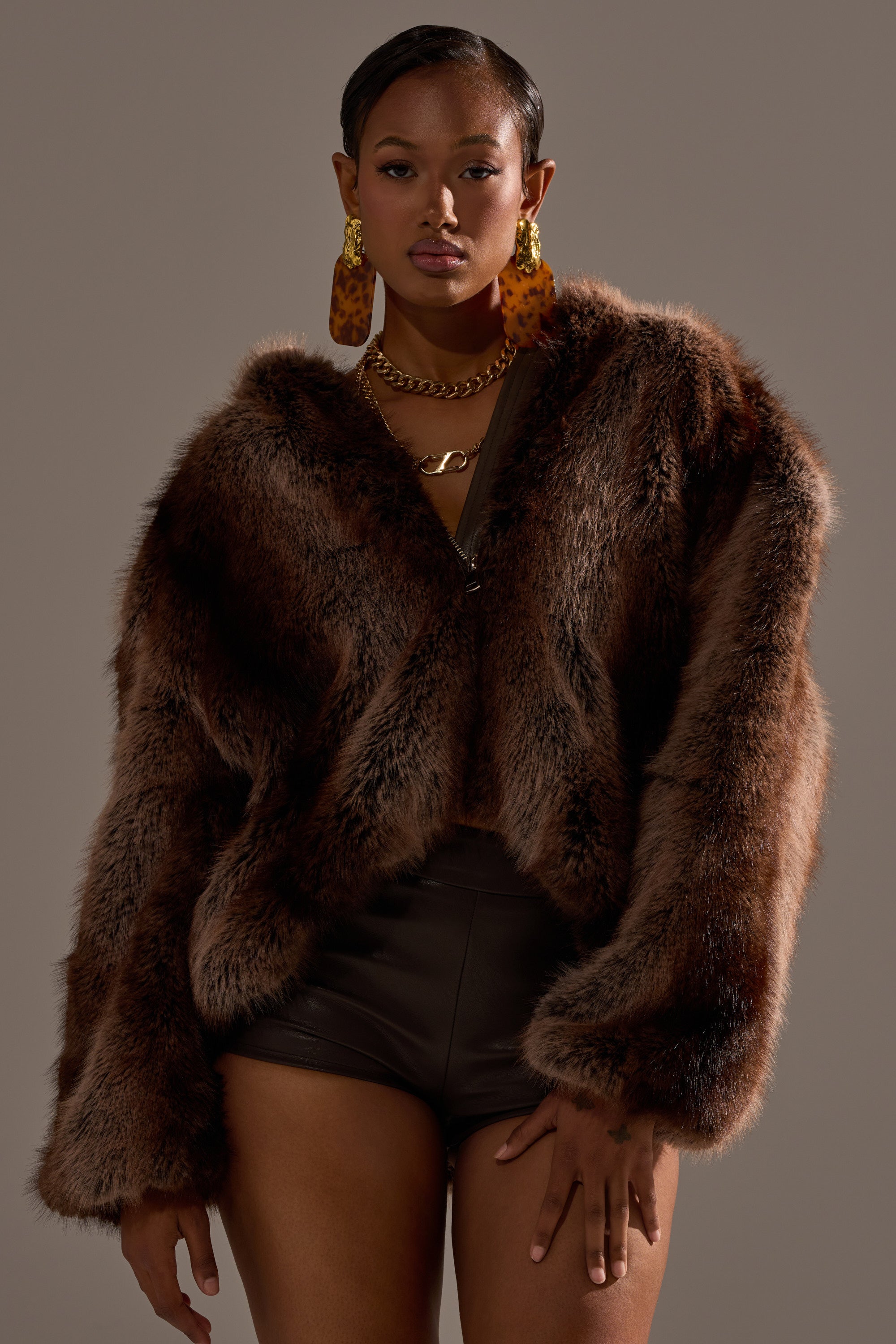 A woman stands confidently in the NATURAL INSTINCTS HOODED FUR BOMBER, styled with dark shorts, large tortoiseshell earrings, and a chunky gold necklace against a plain, neutral background.