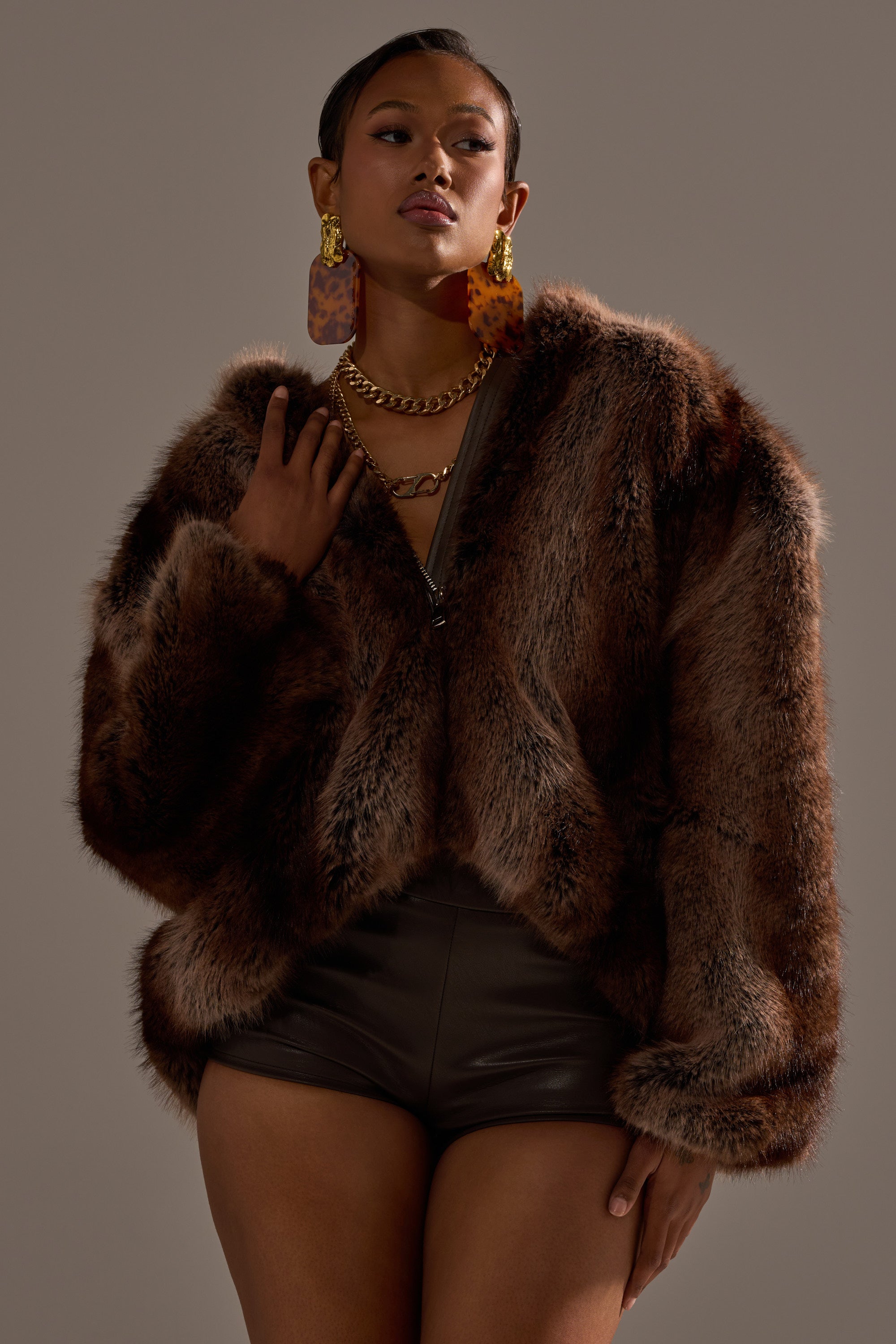A person stands confidently in the NATURAL INSTINCTS HOODED FUR BOMBER with black leather shorts, a chunky gold necklace, and bold statement earrings, posed against a simple neutral background.