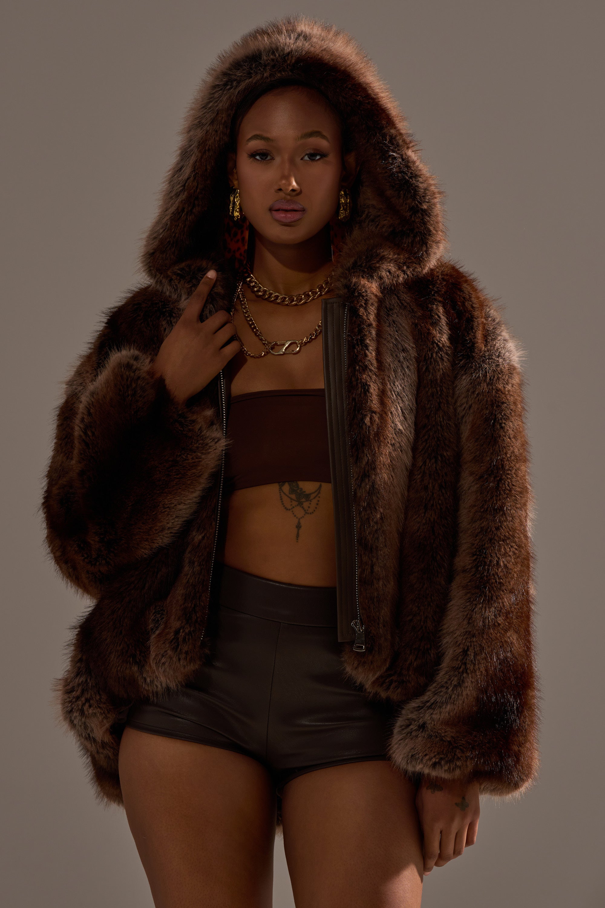 A woman models the NATURAL INSTINCTS HOODED FUR BOMBER over a brown crop top and high-waisted shorts, accessorized with gold jewelry, posing confidently against a neutral background.
