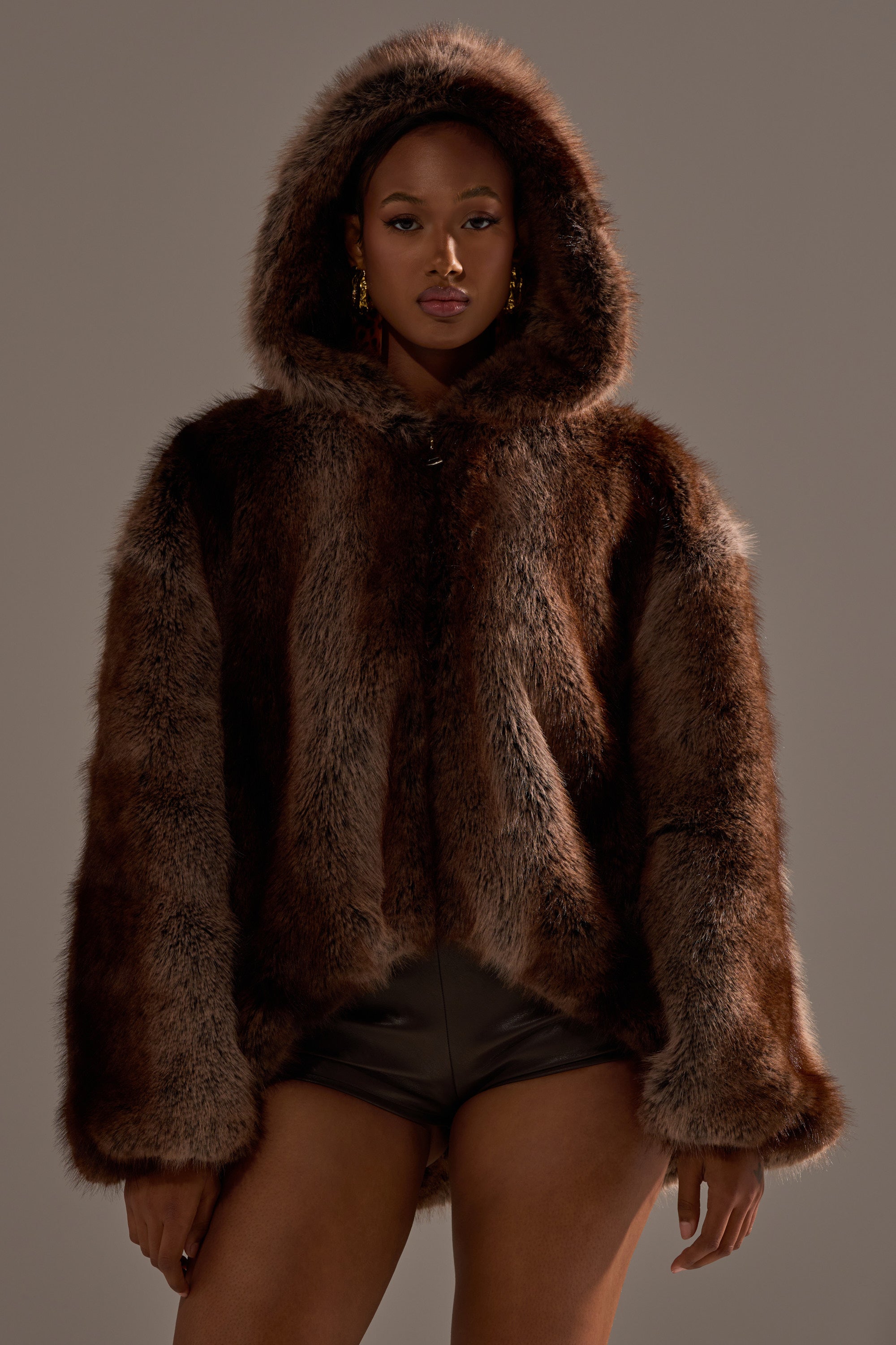 A woman stands confidently against a neutral background, wearing the NATURAL INSTINCTS HOODED FUR BOMBER in brown faux fur paired with black shorts.