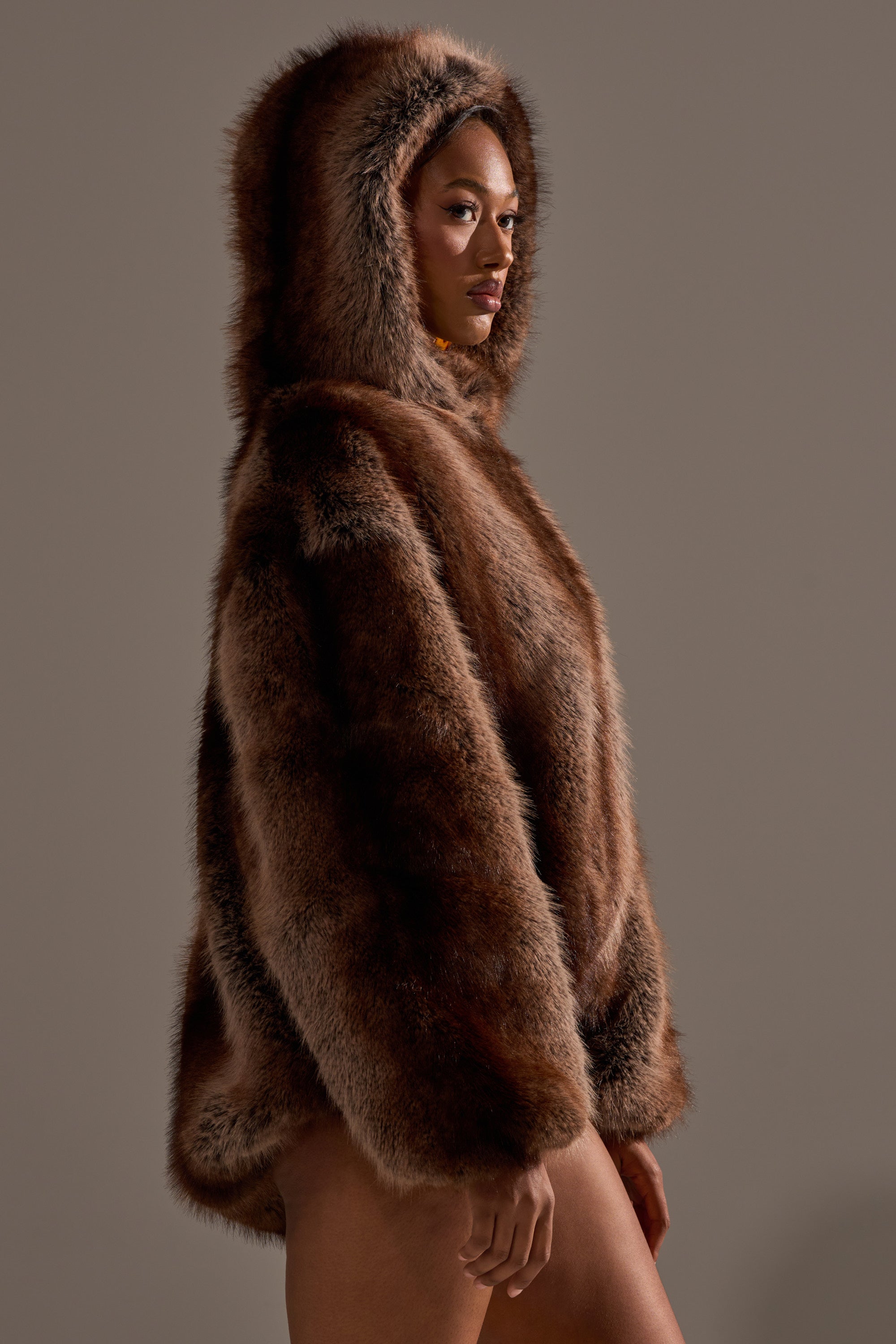 A person stands in profile against a plain background, wearing the NATURAL INSTINCTS HOODED FUR BOMBER—an oversized winter coat with a fur-trimmed hood—hands relaxed by their sides.