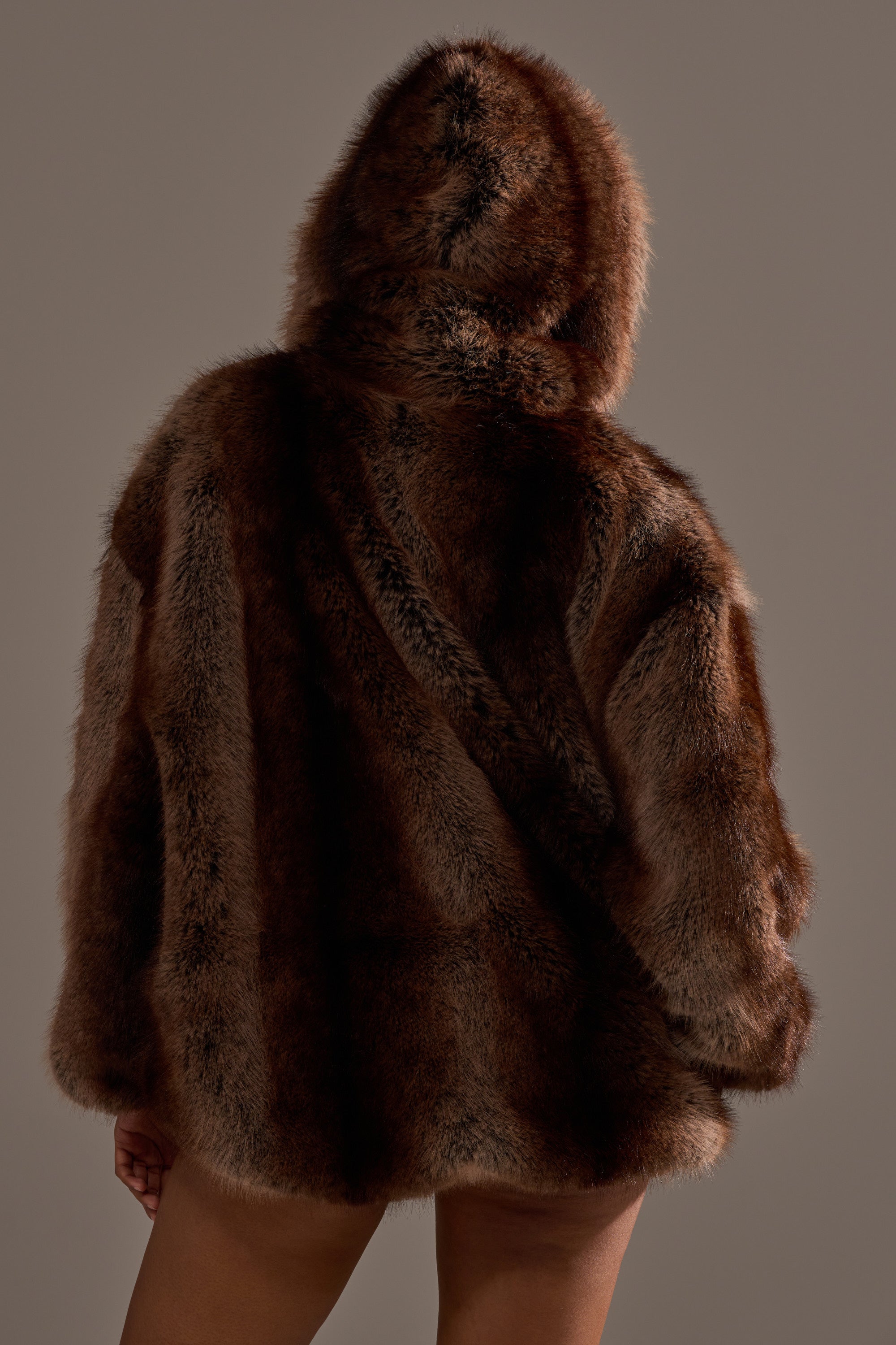 A person is shown from behind wearing the NATURAL INSTINCTS HOODED FUR BOMBER, an oversized winter coat with a faux fur hood, against a plain background. Their legs are visible, but their face is hidden.