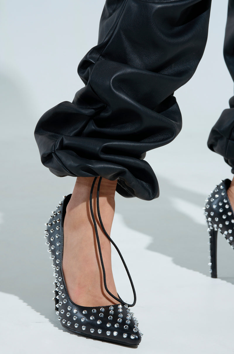A person is dressed in the Charli Stacked PU Jumpsuit in black, featuring pants that gather at the ankles. They wear high-heeled shoes with thin lace straps and metallic studs for added flair.
