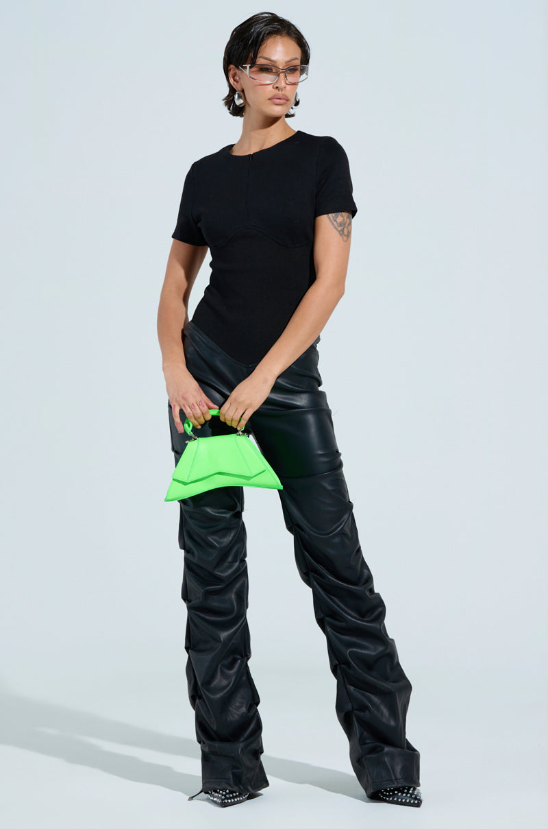 A person wearing the Charli Stacked PU Jumpsuit in black stands confidently, accessorized with stylish sunglasses, a bright green handbag, and pointed studded shoes. The background features a neutral light color.