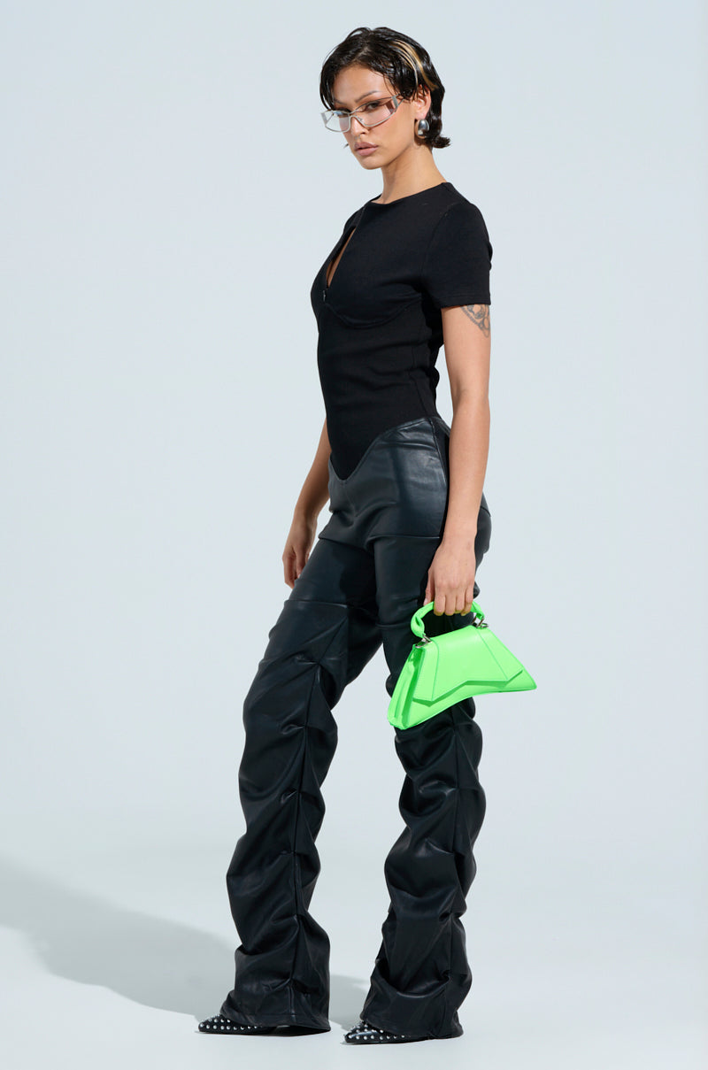 A person stands against a plain background, wearing the CHARLI STACKED PU JUMPSUIT IN BLACK and faux leather pants. They hold a bright green triangular handbag and sport transparent glasses, exuding confidence and a fashion-forward vibe.