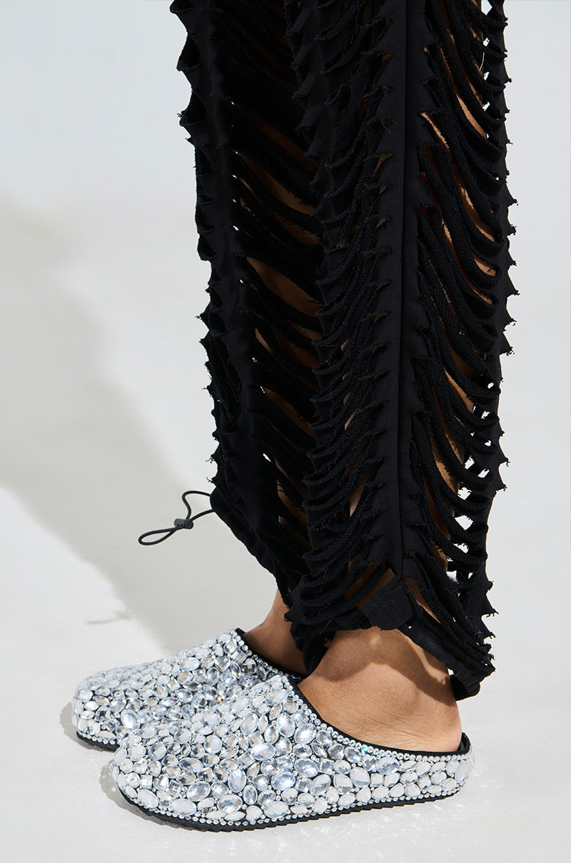 A person wears black pants with vertical shredded slits, revealing an underlayer. Their ensemble is completed with the AZALEA WANG WORLDCLASS EMBELLISHED SLIDE, adorned with shiny translucent crystals.