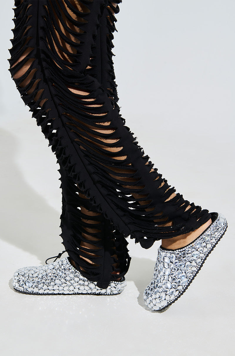 A person in ripped black faux leather pants and AZALEA WANG WORLDCLASS EMBELLISHED SLIDES with glittering gemstones strides across a white surface.