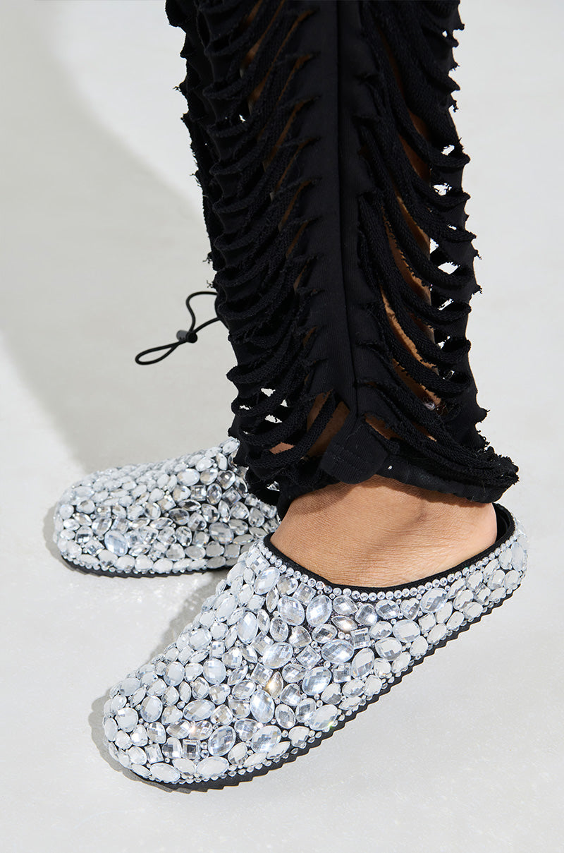 A person wearing intricate black pants with cut-out designs and the AZALEA WANG WORLDCLASS EMBELLISHED SLIDES, encrusted with large, glittering gemstones, stands on a plain surface.