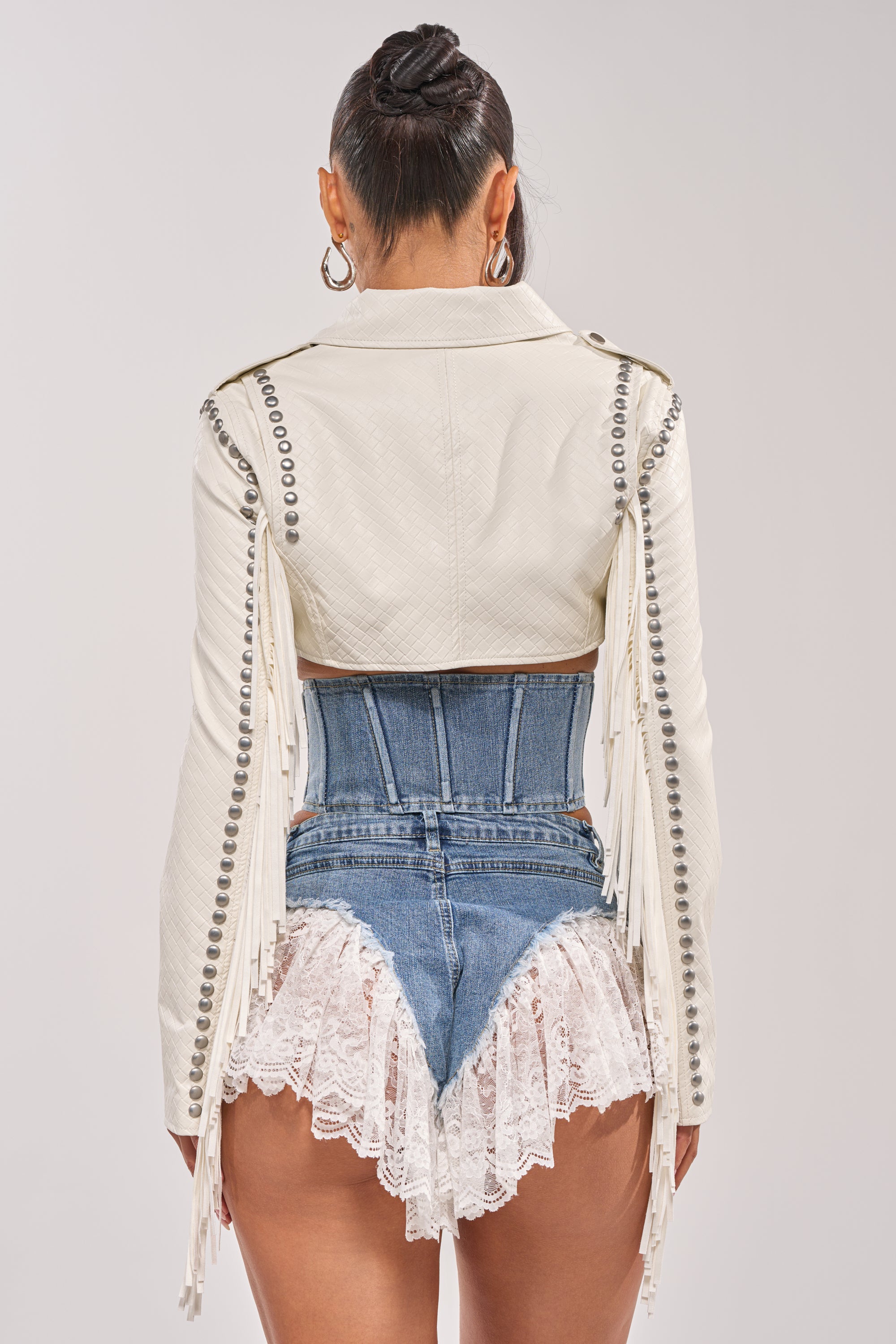 A person stands facing away, wearing the RYDER WOVEN FRINGE MOTO— a Western-inspired white jacket with metallic studs and fringe—paired with denim high-waisted shorts featuring a corset waistband and lace trim, hair in a high bun. 9