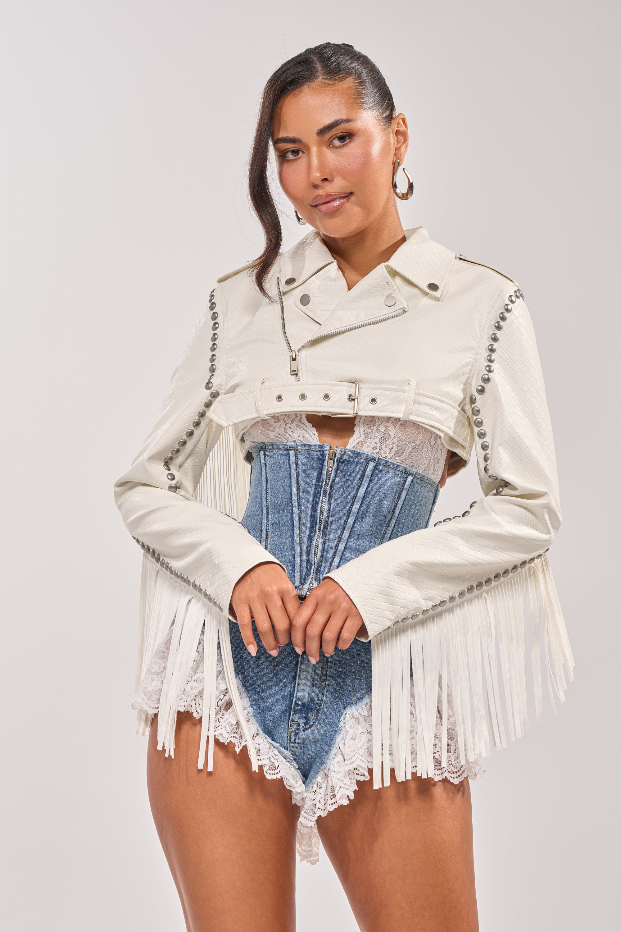 A woman models the RYDER WOVEN FRINGE MOTO, a white faux leather jacket with silver buttons and western fringe, layered over a blue denim corset and white lace bodysuit. She accessorizes with hoop earrings and poses against a light background. 6