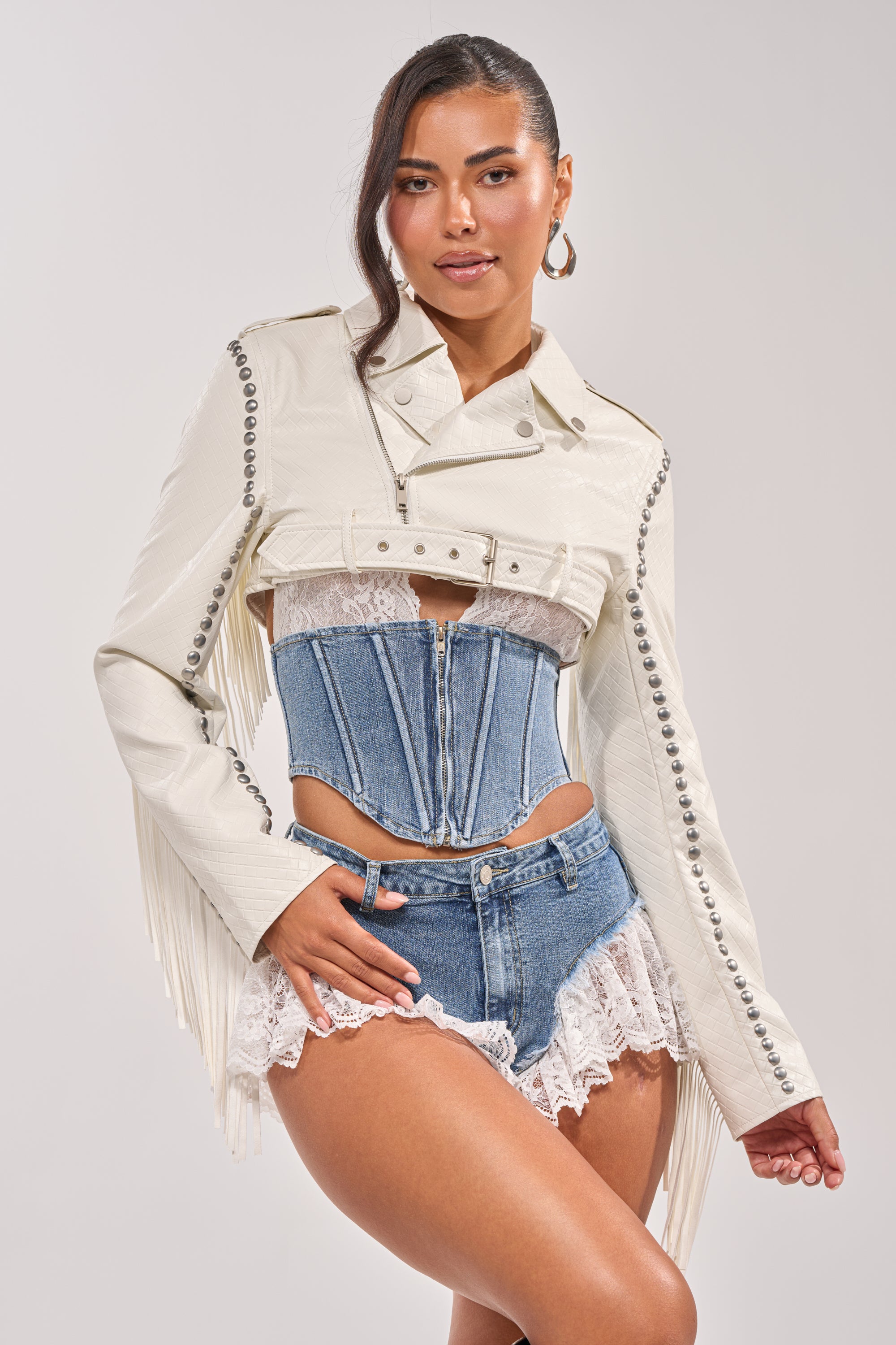A woman poses confidently in the RYDER WOVEN FRINGE MOTO, a cropped white faux leather jacket with silver buttons and fringe, styled with a lace-trimmed denim corset, matching lace-edged denim shorts, hoop earrings, and a side ponytail. 7