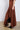 A person stands against a plain background, donning the FOR A LIFETIME MIDI SKIRT from the AKIRA Label. The long, asymmetrical brown skirt flows down to their ankles, revealing fancy brown pointed boots with metal embellishments. Made from stretchy ribbed knit fabric, the skirt features a zipper detail in the front and hugs the upper portion of their body.