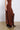 A person is wearing the FOR A LIFETIME MIDI SKIRT from the AKIRA Label, a long, asymmetrical brown skirt that flows past the knees. They have paired it with dark brown leather high-heeled boots embellished with studs and straps. The outfit, featuring a stretchy ribbed knit fabric, stands out against a polished concrete floor and plain white background.