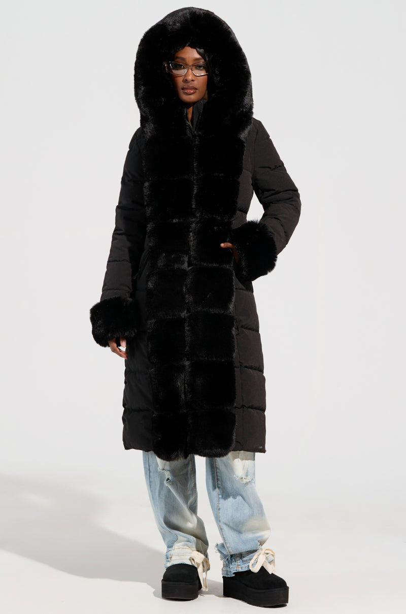 Dressed for a winter adventure, a person puts on the ARLETTE FUR TRIM PUFFER—featuring a cozy faux fur-trimmed hood and cuffs—and pairs it with ripped jeans and black platform shoes, hands tucked in pockets as they stand against a plain background.
