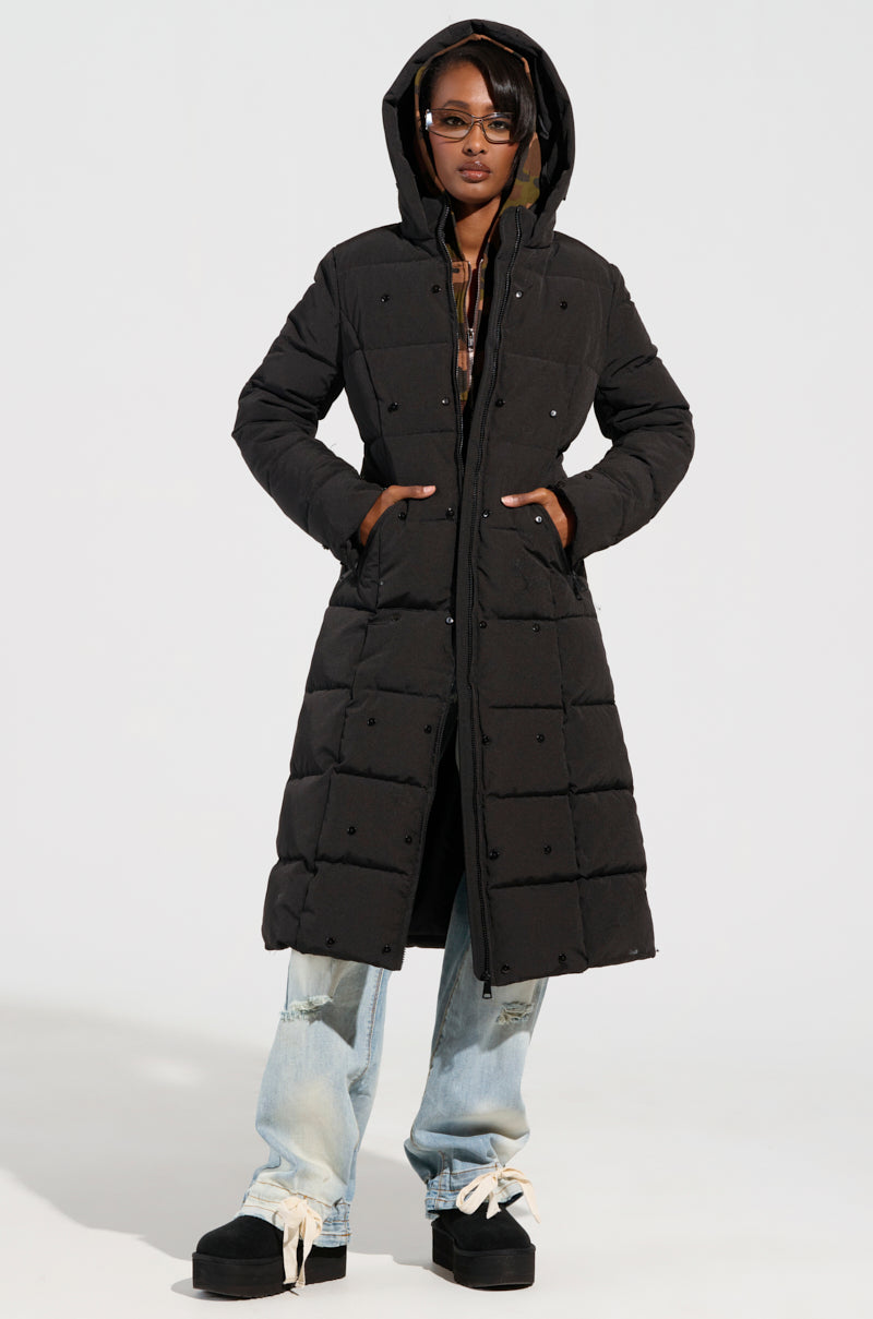 A person stands confidently in the ARLETTE FUR TRIM PUFFER, a long black puffer jacket with faux fur trim. They wear sunglasses and have their hands tucked into the coat pockets, paired perfectly with light blue jeans and black platform shoes. A plain white background sets the scene for this winter adventure.