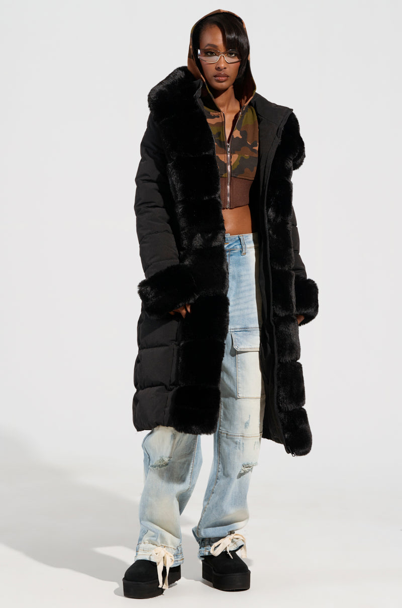 Dressed in the ARLETTE FUR TRIM PUFFER, a person stands confidently over cargo jeans. Beneath, they showcase a camo-patterned cropped jacket that perfectly embodies the spirit of winter adventure. Platform shoes round out this striking ensemble against a plain backdrop.