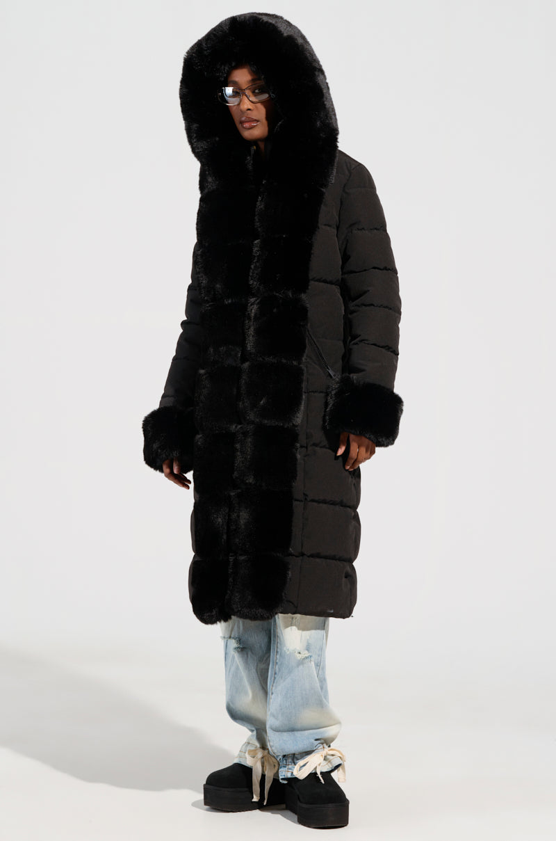 Wearing the ARLETTE FUR TRIM PUFFER, a person stands confidently in a long, black coat adorned with a faux fur hood. Paired with light blue ripped jeans and chunky black shoes, they pose against a plain white background, gearing up for their winter adventure.