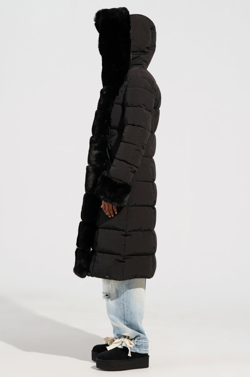 A person stands in profile against a plain background, wearing the ARLETTE FUR TRIM PUFFER, a long black jacket featuring a faux fur trim hood and cuffs. They are paired with light blue jeans and black platform shoes with white laces, perfectly prepared for their winter adventure.