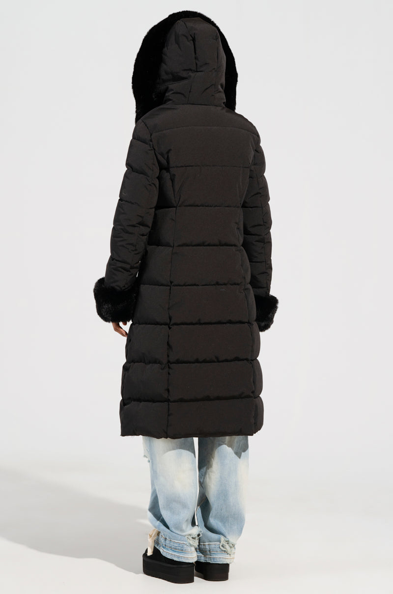 Wearing the ARLETTE FUR TRIM PUFFER, a person stands facing away in a long black puffer jacket featuring faux fur accents on the hood and cuffs. They pair it with light blue jeans and black platform shoes, prepared for a winter adventure against a plain background.