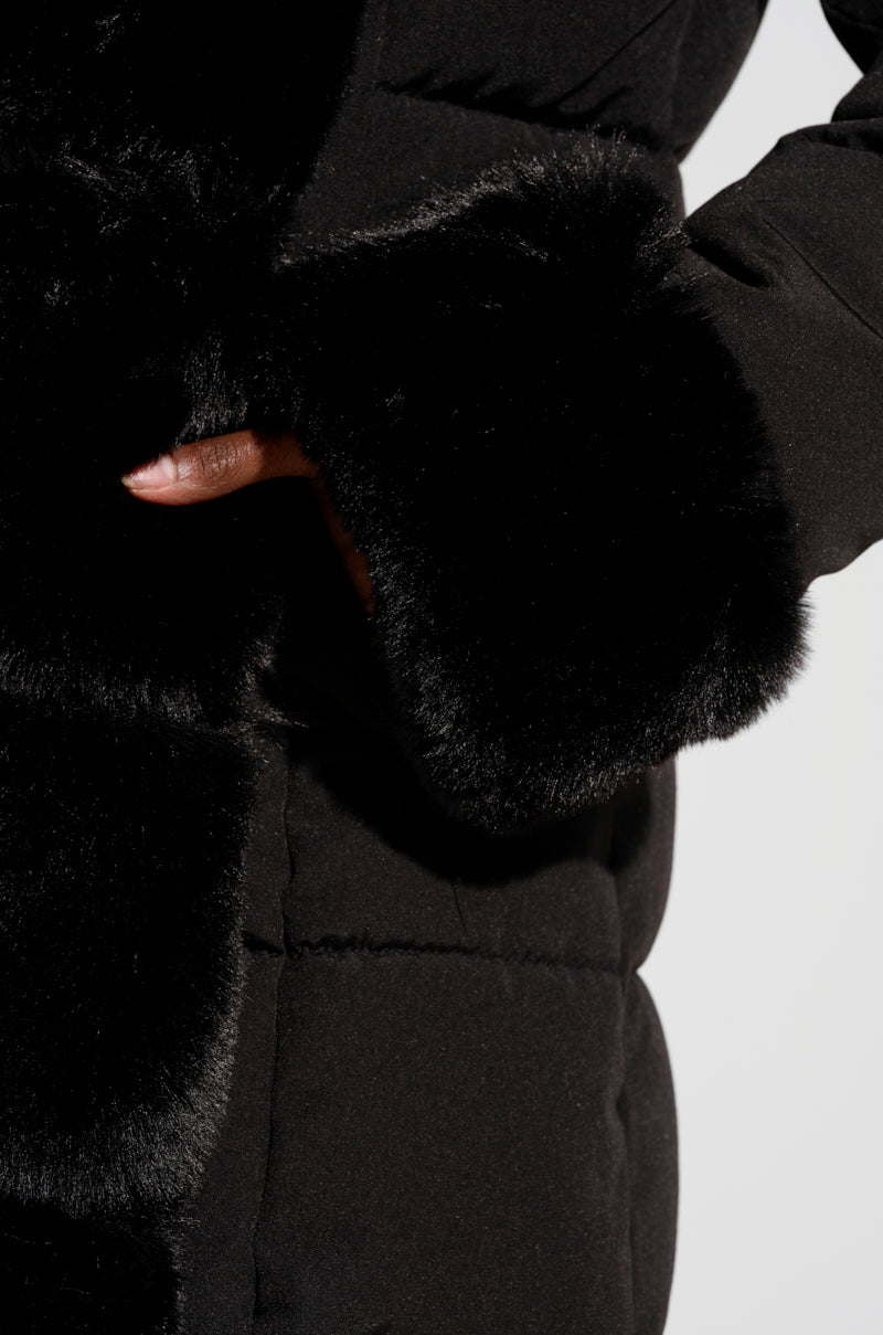 The ARLETTE FUR TRIM PUFFER jacket in black features faux fur trim on the cuffs and front edge, revealing a hand partially tucked inside. The fabric appears soft, offering a luxurious texture that's ideal for any winter adventure.