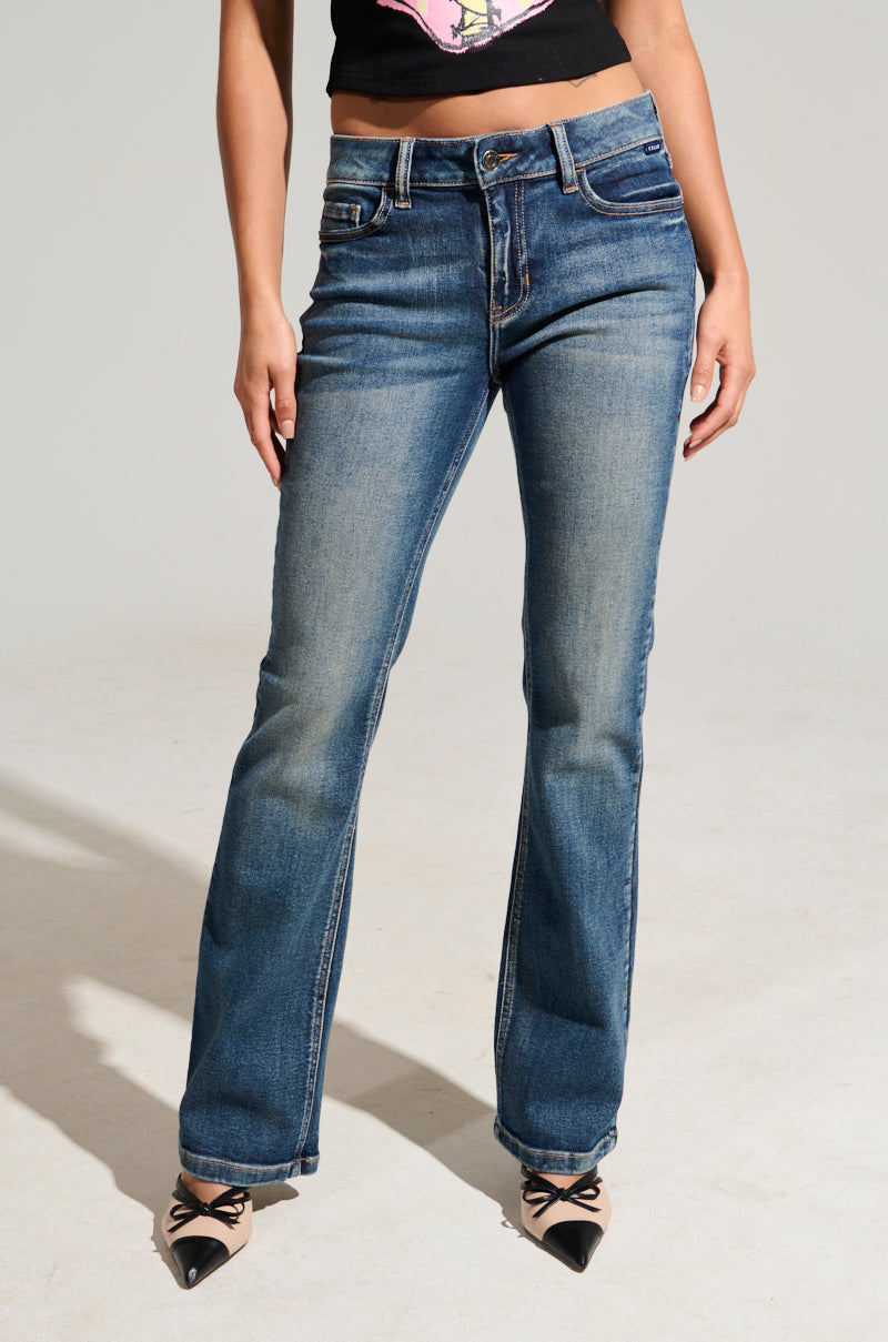 A person wearing the Fine Fine Baby Denim Pant, crafted from semi-stretchy denim in a faded blue with a flared leg silhouette, and accompanied by a black top stands against a plain background. The jeans include front pockets, while the pointed-toe shoes have bow details. The image does not show the person's head.