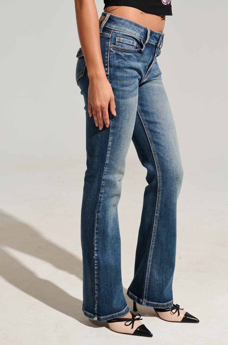 A person wearing FINE FINE BABY DENIM PANT, with a flared style crafted from semi-stretchy blue denim, and black pointed-toe ballet flats stands on a light gray floor. The individual is posed with one arm relaxed at their side, partially revealing their top.