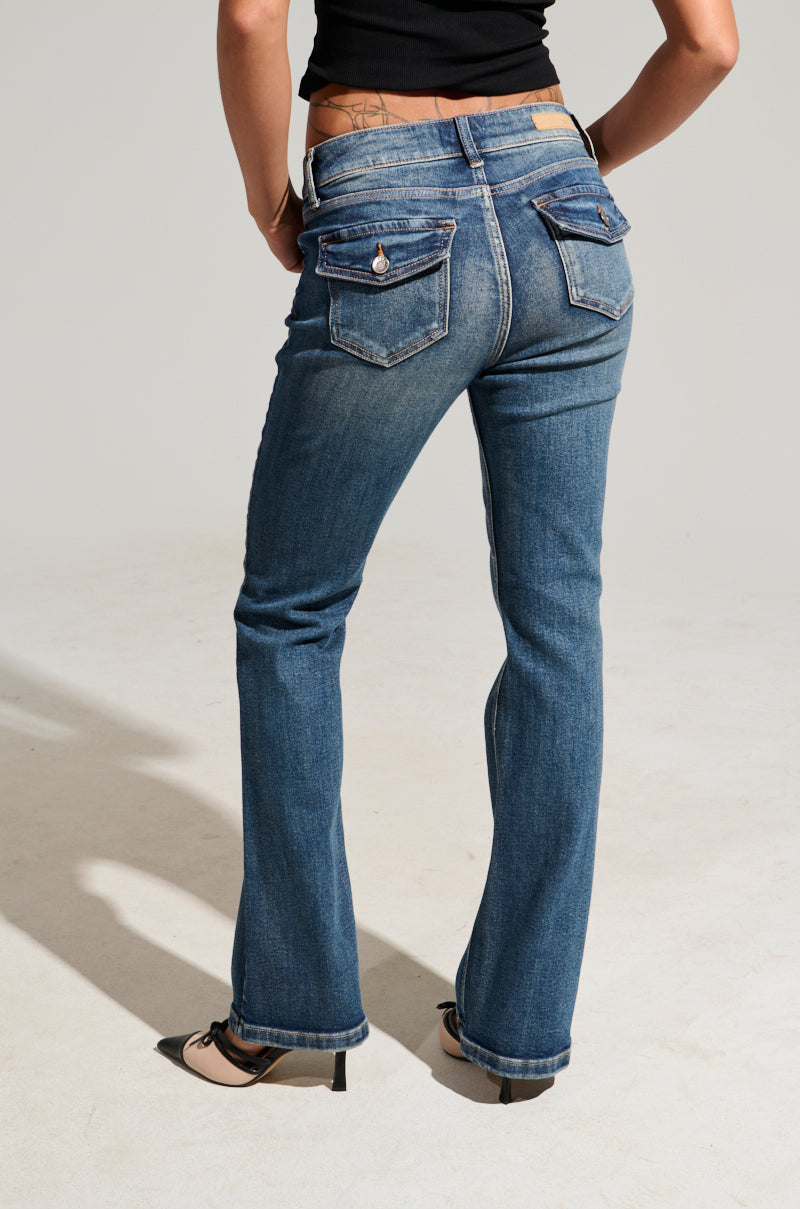 A person wearing the FINE FINE BABY DENIM PANT stands against a neutral background, showcasing its semi-stretchy denim fabric in blue with a subtle flared leg silhouette. The jeans are adorned with decorative pockets and slight fading. The individual completes the chic ensemble with a black top and black heels.