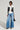 Against a plain backdrop, a person exudes style in a black and white leather jacket paired with a white top featuring a cutout. They sport the GET TO IT DENIM PANT in a high-rise, wide-leg silhouette, matched with black pointed heels and a small black purse.
