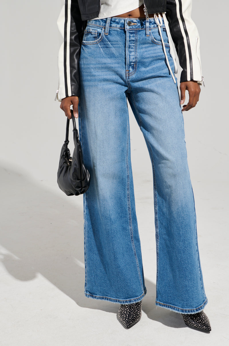 A person in a black and white jacket complements their high-rise, wide-leg silhouette with the GET TO IT DENIM PANT, clutching a small black handbag. Their pointed, textured shoes add a stylish touch against the plain, light-colored background.