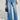 A person in a black and white jacket complements their high-rise, wide-leg silhouette with the GET TO IT DENIM PANT, clutching a small black handbag. Their pointed, textured shoes add a stylish touch against the plain, light-colored background.