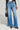 A person in a black and white jacket complements their high-rise, wide-leg silhouette with the GET TO IT DENIM PANT, clutching a small black handbag. Their pointed, textured shoes add a stylish touch against the plain, light-colored background.