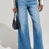 A person in a black and white jacket complements their high-rise, wide-leg silhouette with the GET TO IT DENIM PANT, clutching a small black handbag. Their pointed, textured shoes add a stylish touch against the plain, light-colored background.