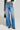 A person wearing the "GET TO IT DENIM PANT" with a high-waisted and wide-leg silhouette, paired with a black and white striped jacket, stands holding a small black purse. They are also wearing pointed, patterned shoes against a neutral gray background.