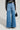 A person wearing the GET TO IT DENIM PANT, a black and white jacket, and holding a small black bag stands with their back to the camera against a plain background.