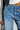A person is seen wearing the GET TO IT DENIM PANT, featuring a wide leg silhouette in blue denim, paired with a black and white jacket. They are holding a small black bag. The close-up highlights the high-rise design of the jeans and part of the jacket, emphasizing the casual outfit style.