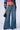 Person wearing the ONE MORE WEEKEND FLARE DENIM PANT and a colorful, striped cropped top. They are holding a small black handbag, and black shoes are partially visible at the bottom of the pants. The background is plain and white.