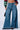 A person is wearing the ONE MORE WEEKEND FLARE DENIM PANT, featuring high-waisted, wide-leg blue jeans with distressed detailing and their hands in their pockets. The top is partially visible, revealing a colorful, long-sleeved crop top. They are carrying a small black handbag and wearing black shoes.