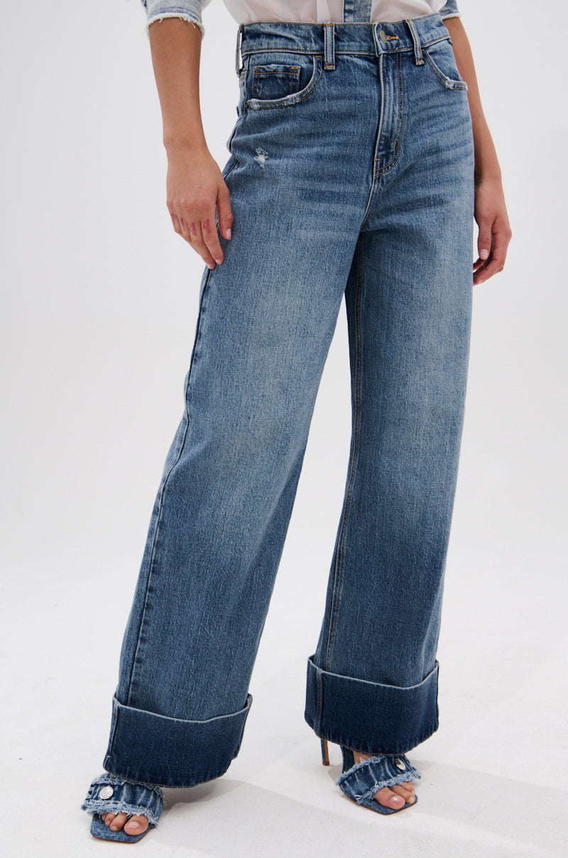 A person wears JORDAN WIDE LEG DENIM with a high-rise, wide-leg silhouette, large cuffed hems, and distressed details, paired with open-toe heeled shoes. The photo is cropped at the shoulders.