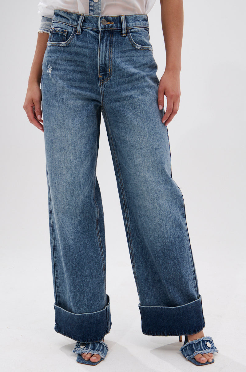 Someone wears the JORDAN WIDE LEG DENIM, featuring a high-rise, wide-leg silhouette in blue with large cuffed hems and a slightly distressed look, paired with a white shirt and open-toed denim heels against a plain white background.