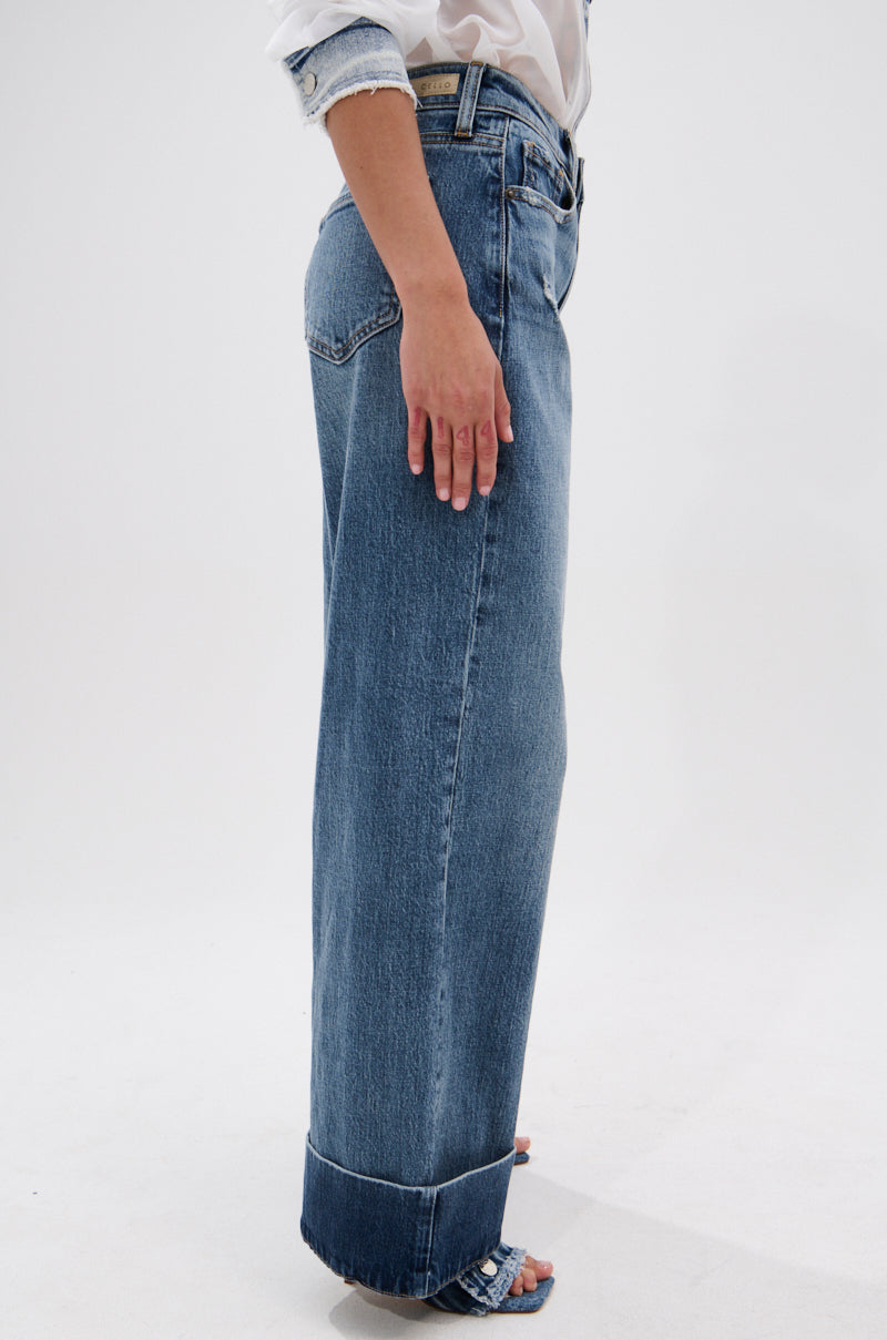 A person stands sideways against a plain white background, wearing JORDAN WIDE LEG DENIM with a high-rise, wide-leg fit and cuffed hem, paired with a white shirt.