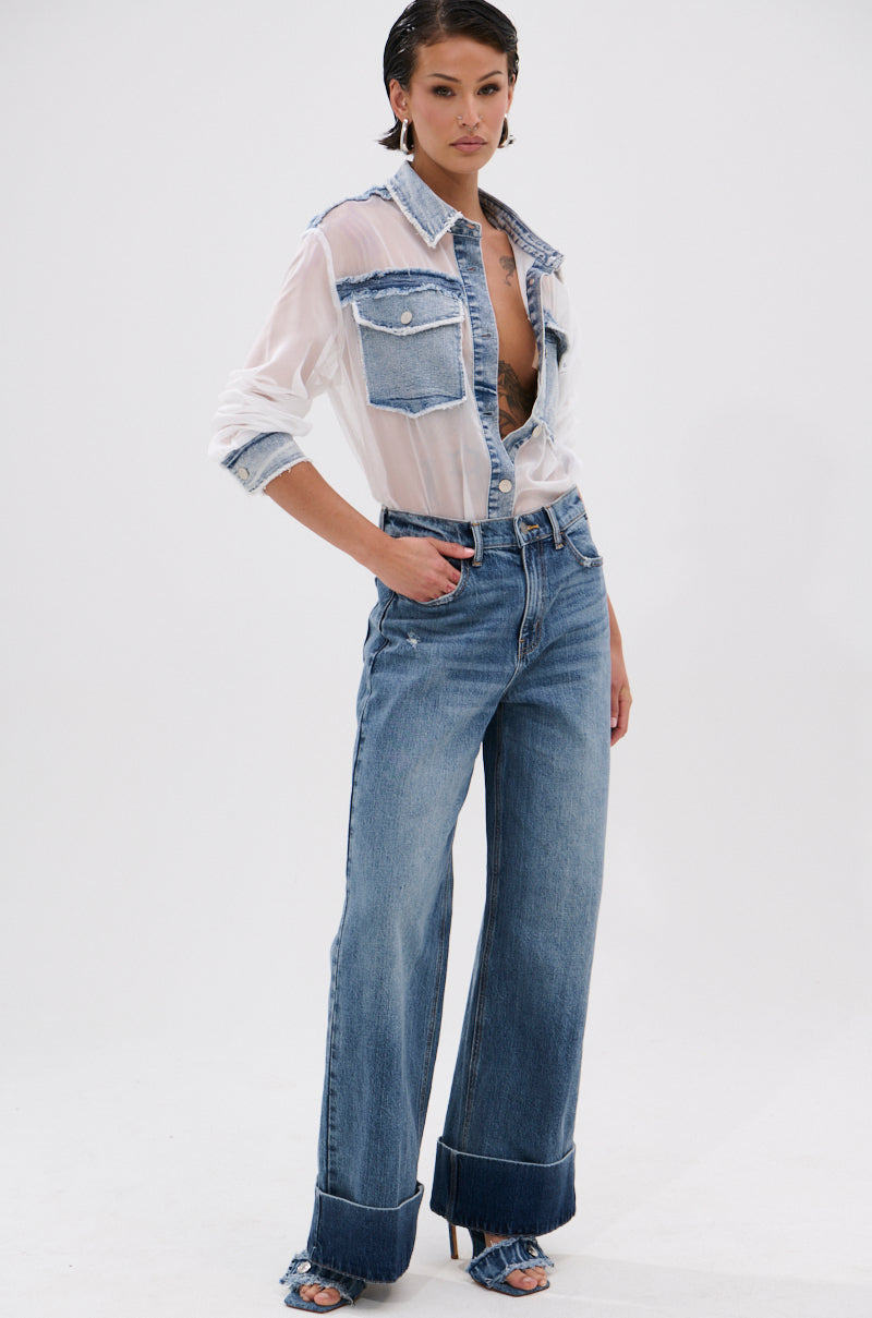 A woman poses confidently in JORDAN WIDE LEG DENIM—high-rise, wide-leg jeans in medium-blue, cuffed at the ankles—styled with a sheer white button-down over a bralette and open-toe heels against a plain white background.